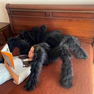 May include: A large black fuzzy spider plush toy on a bed with a person reading a book. The spider is laying on the bed with its legs spread out. The person is lying on their side with their head resting on the spider's body.