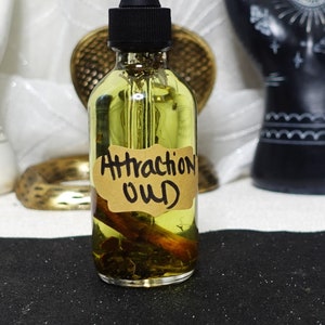 May include: A clear glass bottle with a black dropper top and a brown label that reads "Attraction Oud". The bottle is filled with a yellow liquid and contains a cinnamon stick. The bottle is sitting on a black surface.