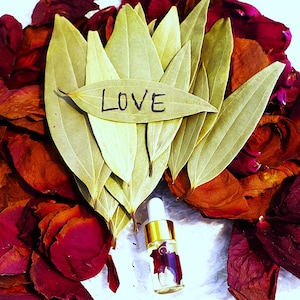 Może przedstawiać: Suszone płatki róż w odcieniach czerwieni i pomarańczy otaczają małą szklaną butelkę ze złotą zakrętką. Butelka zawiera przezroczysty płyn, a na zielonym liściu napisano słowo "LOVE".