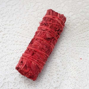 May include: A bundle of red sage, tightly wrapped with red twine. The sage has a textured appearance, and the twine is wrapped around the bundle multiple times. The background is a white, patterned surface.