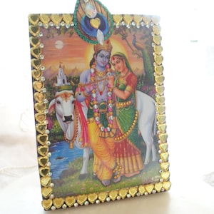 May include: A framed print depicting Hindu deities, Radha and Krishna, with a cow and peacock. The frame is decorated with gold heart-shaped embellishments and a peacock feather. The print features vibrant colors and intricate details.