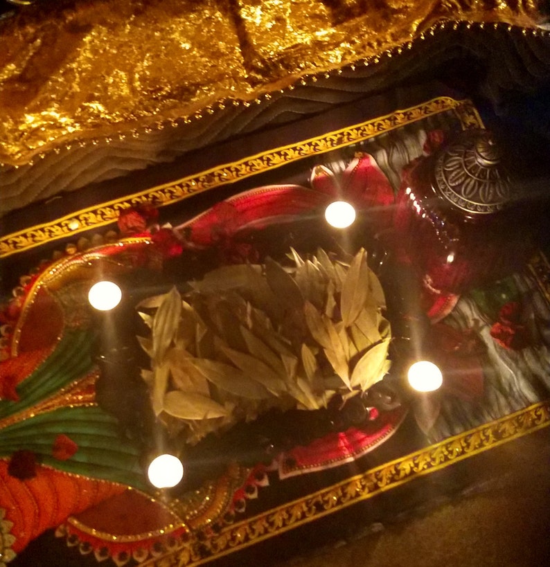 K&ouml;nnte beinhalten: Ein schwarzer Teppich mit goldenen Mustern und einem Haufen getrockneter Bl&auml;tter in der Mitte. Vier brennende Kerzen umgeben die Bl&auml;tter, und ein kleiner, verzierter, rot-goldener Beh&auml;lter befindet sich rechts von den Bl&auml;ttern.