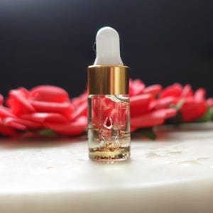 Może przedstawiać: Mała szklana butelka z zakraplaczem ze złotą nasadką i białą końcówką zakraplacza. Butelka zawiera przezroczysty płyn z małymi cząsteczkami. Czerwone róże są rozmyte w tle, z białą powierzchnią.