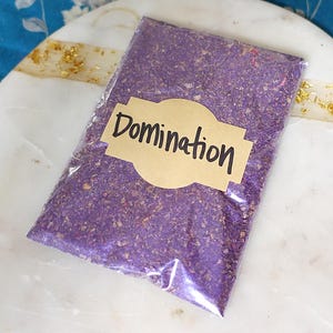 May include: A rectangular clear plastic pouch filled with a coarse purple granular substance. A tan paper label is adhered to the front of the pouch with the word Domination printed in black ink in a stylized, handwritten-style font.