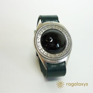 Peut inclure: Une montre-bracelet unique avec un grand cadran noir arrondi serti dans un boîtier en métal argenté. La montre est dotée d'un bracelet en cuir vert foncé et d'une lunette marquée de chiffres. La marque "ragalaxys" est visible en bas.