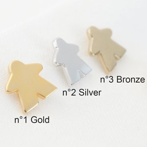 Op de afbeelding: Drie metalen spelstukken in goud, zilver en brons. De stukken zijn gevormd als een persoon en bevinden zich op een witte achtergrond. De tekst 'n°1 Goud', 'n°2 Zilver' en 'n°3 Brons' staat onder de stukken.