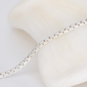 Puede incluir: Un collar de cadena de plata con estilo de cadena de caja. La cadena está sobre una superficie blanca.