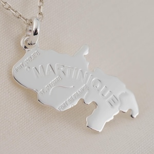 Puede incluir: Un collar con colgante plateado que representa la isla de Martinica. El colgante es un contorno detallado de la isla, con la palabra "MARTINIQUE" grabada. El collar cuelga de una fina cadena de plata.