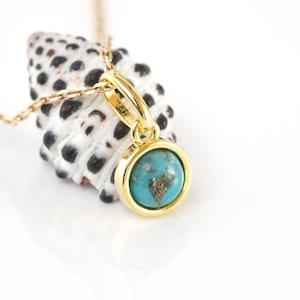 Puede incluir: Un collar de cadena de oro con un colgante de piedra turquesa. El colgante está engastado en un bisel de oro y tiene una forma redonda. El collar está apoyado sobre una concha.