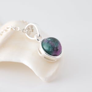 Op de afbeelding: Een zilveren hangerketting met een ronde edelsteen met groene en paarse tinten. De hanger is gezet in een zilveren bezel en hangt aan een fijne zilveren ketting. De ketting wordt gepresenteerd op een wit oppervlak.