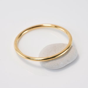Peut inclure: Une fine bague dorée repose sur une petite pierre grise claire. La bague est circulaire et semble être en métal. Le fond est une surface blanche unie, offrant une présentation épurée et simple du bijou.