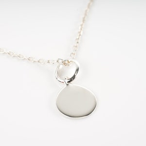 Puede incluir: Un collar de cadena de plata con un colgante redondo y liso. El colgante está unido a la cadena con un pequeño lazo de plata.