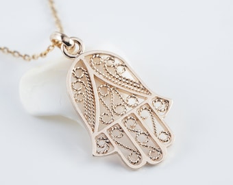 May include: A gold chain necklace with a delicate, filigree hamsa hand pendant. The pendant is intricately designed with a detailed pattern.