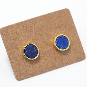 Pode incluir: Um par de brincos de pressão de ouro com pedras de lápis-lazúli azuis. Os brincos são exibidos em um cartão marrom.