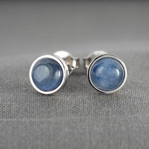 Op de afbeelding: Een paar oorknopjes met ronde, blauwe edelstenen gezet in zilveren bezels. De edelstenen hebben een gemarmerd uiterlijk en de oorbellen worden getoond tegen een grijze achtergrond. Sieraden.
