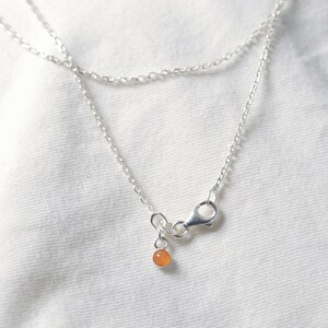 May include: A silver chain necklace with a small orange gemstone pendant. The necklace has a lobster clasp closure.