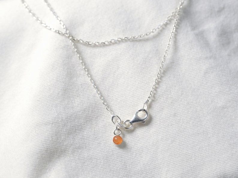 May include: A silver chain necklace with a small orange gemstone pendant. The necklace has a lobster clasp closure.