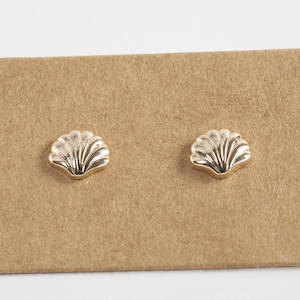 以下が含まれることがあります： 茶色のカードの上に置かれた、金色の貝殻のスタッドピアスのペア