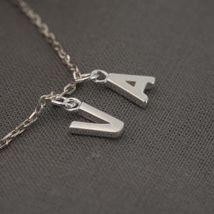 Peut inclure: Un collier en chaîne argent avec deux breloques argentées, l'une avec la lettre 'V' et l'autre avec la lettre 'A'.