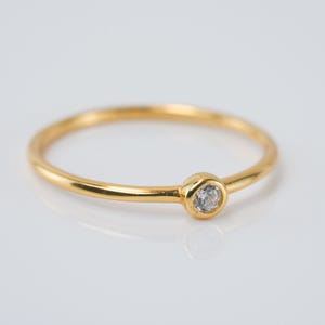 Puede incluir: Un delicado anillo de oro con una pequeña gema redonda y transparente engastada en un bisel. La banda del anillo es fina y pulida, reflejando la luz. El anillo está sobre un fondo blanco.