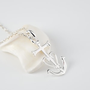 Op de afbeelding: Een zilveren ketting met een hanger met een kruis en een anker die in elkaar zijn gevlochten. De hanger ligt op een witte achtergrond.