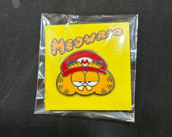 Puede incluir: Un pin de esmalte amarillo y naranja con un gato de dibujos animados que lleva un gorro de Mario rojo y blanco. El pin dice "MEOWRIO" en una burbuja amarilla.