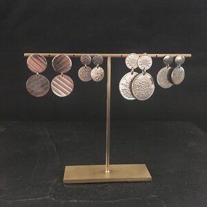 Peut inclure: Trois paires de boucles d'oreilles en argent pr&eacute;sent&eacute;es sur un support dor&eacute;. Les boucles d'oreilles sont toutes rondes et ont des motifs diff&eacute;rents. Les boucles d'oreilles de gauche ont une surface textur&eacute;e, les boucles d'oreilles du milieu ont un motif floral et les boucles d'oreilles de droite ont un motif circulaire.