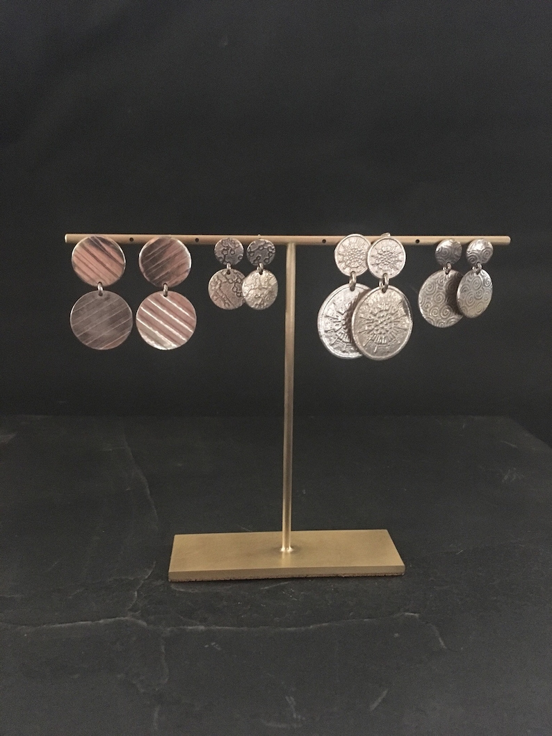 Peut inclure: Trois paires de boucles d'oreilles en argent pr&eacute;sent&eacute;es sur un support dor&eacute;. Les boucles d'oreilles sont toutes rondes et ont des motifs diff&eacute;rents. Les boucles d'oreilles de gauche ont une surface textur&eacute;e, les boucles d'oreilles du milieu ont un motif floral et les boucles d'oreilles de droite ont un motif circulaire.