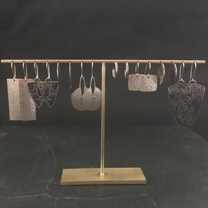 Pode incluir: Uma cole&ccedil;&atilde;o de brincos de prata com v&aacute;rios designs, incluindo padr&otilde;es geom&eacute;tricos, florais e abstratos. Os brincos est&atilde;o pendurados em um suporte dourado sobre um fundo preto.