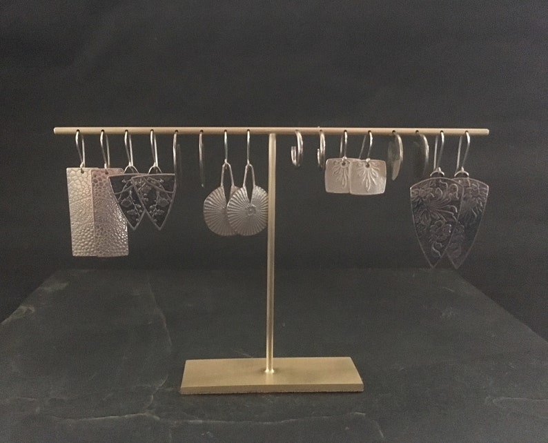 Pode incluir: Uma cole&ccedil;&atilde;o de brincos de prata com v&aacute;rios designs, incluindo padr&otilde;es geom&eacute;tricos, florais e abstratos. Os brincos est&atilde;o pendurados em um suporte dourado sobre um fundo preto.