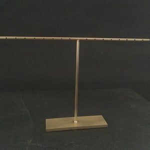 Pode incluir: Um suporte de joias de metal dourado com uma barra horizontal longa e uma base retangular. A barra tem v&aacute;rios orif&iacute;cios para segurar brincos.