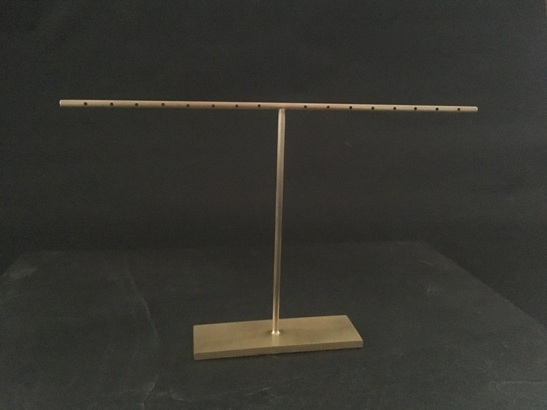 Pode incluir: Um suporte de joias de metal dourado com uma barra horizontal longa e uma base retangular. A barra tem v&aacute;rios orif&iacute;cios para segurar brincos.