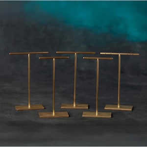 May include: Five gold-colored metal jewelry display stands. Each stand has a rectangular base, a vertical post, and a horizontal bar at the top. The stands are arranged against a dark background with a teal and blue gradient.