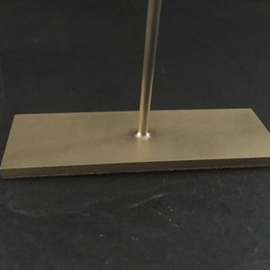Pode incluir: Um suporte de metal dourado com uma base retangular e uma &uacute;nica haste vertical.