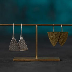 Puede incluir: Dos pares de pendientes expuestos en un soporte dorado. Un par es plateado con un diseño de hojas, el otro es dorado con una superficie texturizada. Los pendientes cuelgan de finos ganchos plateados.