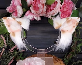May include: A pair of white and tan spotted cat ears attached to a clear headband. The ears have a fluffy texture and are set against a backdrop of pink peonies and greenery. The headband is displayed on a clear stand.