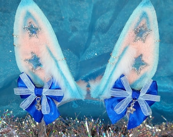 May include: A pair of blue and white faux fur bunny ears with pink inner ears. The ears are decorated with silver star-shaped embellishments and small silver hoops. Each ear has a blue bow with a silver charm. The ears are attached to a clear headband.
