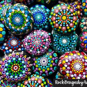 May include: A collection of painted rocks with colorful mandala designs. Each rock is painted with a different pattern of dots and circles in various colors, including pink, blue, green, yellow, and red. The rocks are arranged in a pile, with some overlapping. The text "RockDesignsbyAmanda" is visible at the bottom of the image.