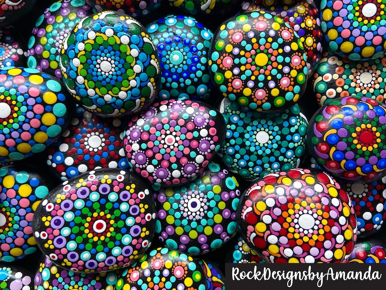May include: A collection of painted rocks with colorful mandala designs. Each rock is painted with a different pattern of dots and circles in various colors, including pink, blue, green, yellow, and red. The rocks are arranged in a pile, with some overlapping. The text "RockDesignsbyAmanda" is visible at the bottom of the image.