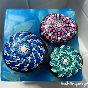 May include: Three painted rocks with mandala designs. The rocks are black with white, blue, pink, and green dots. The rocks are on a blue surface. RockDesignsbyAmanda is written in the bottom right corner.