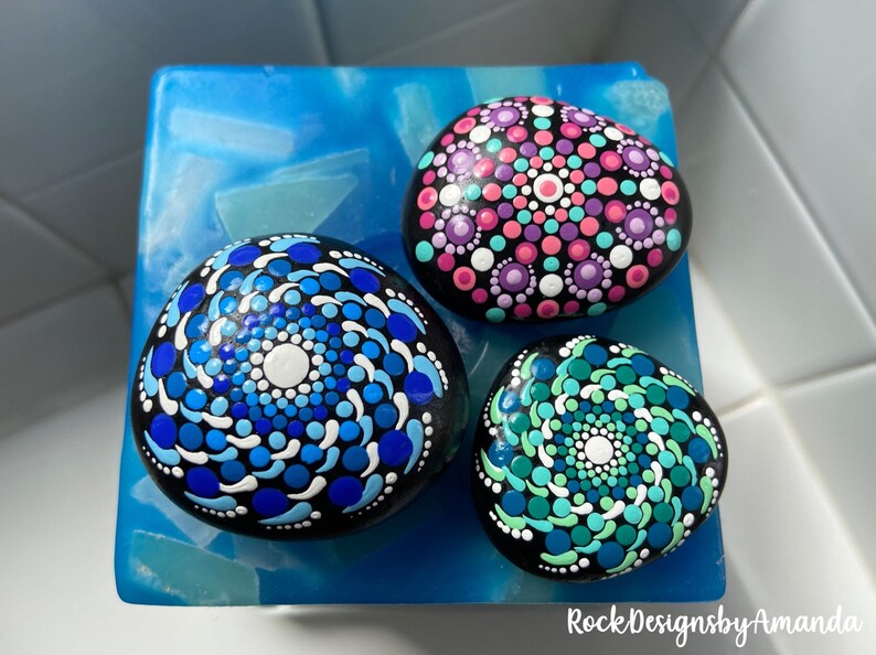 May include: Three painted rocks with mandala designs. The rocks are black with white, blue, pink, and green dots. The rocks are on a blue surface. RockDesignsbyAmanda is written in the bottom right corner.