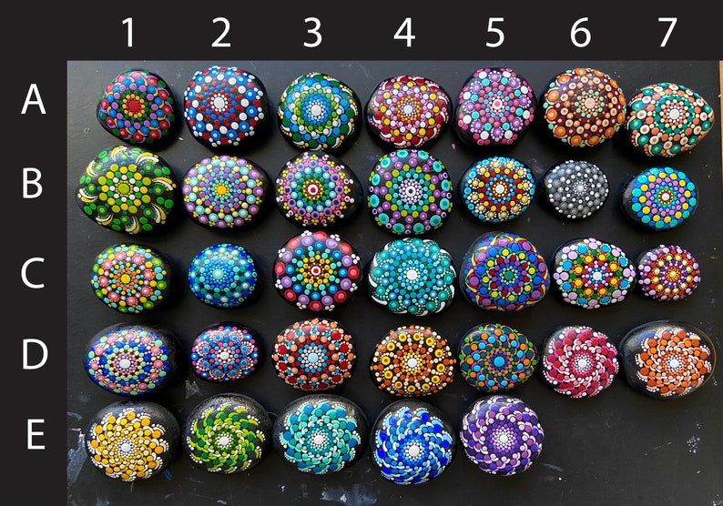 May include: A collection of 35 painted rocks with mandala designs in various colors, arranged in a grid pattern on a black surface. The rocks are labeled with letters A through E and numbers 1 through 7.