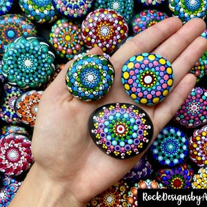 May include: A hand holds three painted rocks with colorful mandala designs. The rocks are surrounded by other painted rocks with similar designs.
