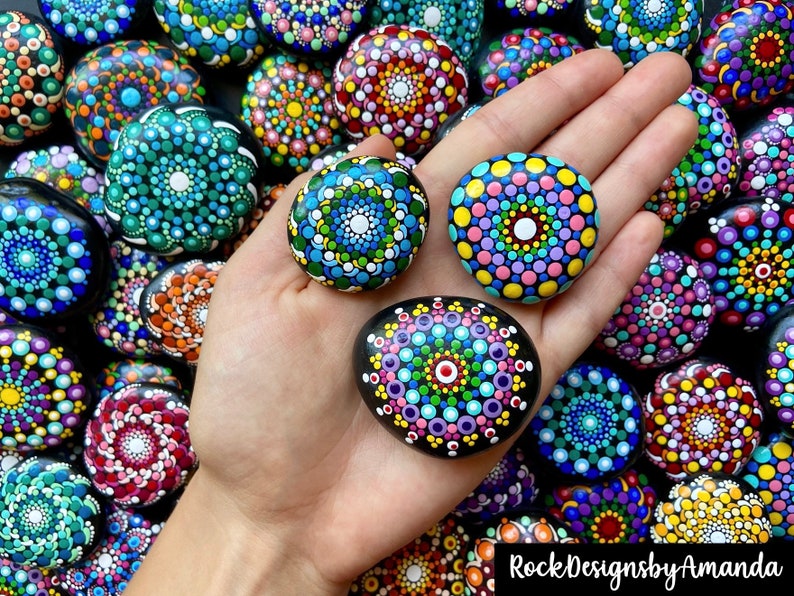 May include: A hand holds three painted rocks with colorful mandala designs. The rocks are surrounded by other painted rocks with similar designs.