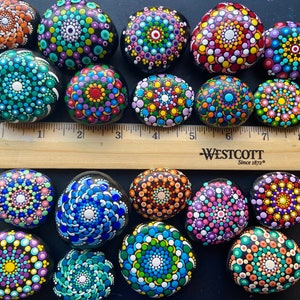 May include: A collection of 17 painted rocks with colorful mandala designs. The rocks are arranged on a black surface with a wooden ruler labeled "Westcott Since 1872" underneath.