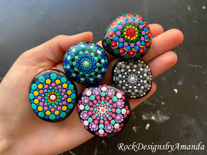 May include: Five painted rocks with mandala designs. The rocks are painted in various colors, including pink, blue, green, yellow, white, black, and gray. The rocks are arranged in a circle on a hand.