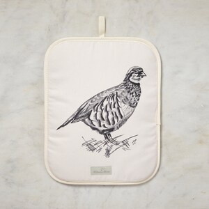 May include: A white cotton pot holder with a black and white illustration of a bird. The bird is a partridge and is standing on a bed of grass. The pot holder has a loop for hanging.