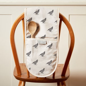May include: A white oven mitt with a black and white pattern of birds. The mitt is hanging on the back of a wooden chair with a wooden spoon inside the pocket.