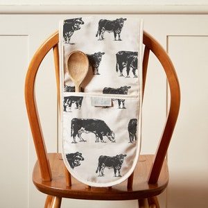 May include: A white oven mitt with a black cow print design. The mitt has a pocket on the side and a wooden spoon is hanging from the pocket.