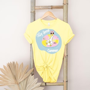 May include: Yellow t-shirt with a graphic of a cartoon goose wearing a pink bow and sunglasses, floating in a pool with the words "Silly Goose Summer". The shirt is knotted at the waist and hangs on a wooden hanger.